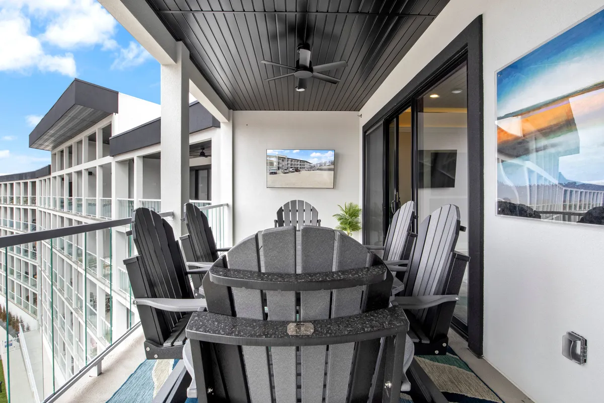 A spacious outdoor balcony featuring an inviting dining table set, Adirondack chairs, a ceiling fan, a wall-mounted TV, and framed artwork. The scene is bright and airy, with an open view suggesting a relaxing atmosphere perfect for group gatherings, al fresco meals, and unwinding after a day of fun.