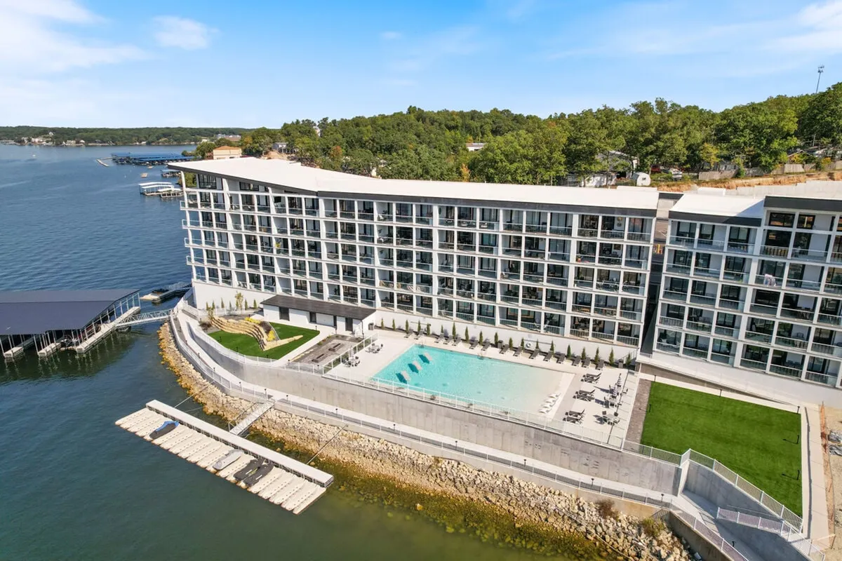 The image showcases the stunning exterior of a modern lakeside condo at sunset, with a sparkling swimming pool in the foreground, manicured green lawn, and the vast lake stretching out behind. The relaxed scene hints at activities like swimming, sunbathing, boating, and evening gatherings, with inviting fire pits and the warm glow of dusk over the water.