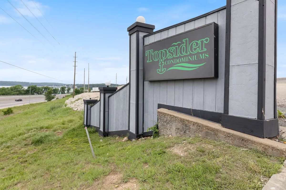 This exterior shot showcases the Topsider Condominiums sign, a staircase leading up to the building, and green grass in a tranquil setting. The composition evokes a sense of arrival, community, and access to nearby outdoor activities.