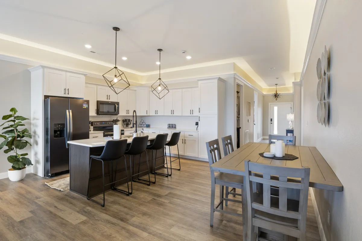 This image shows a spacious, modern kitchen with a large kitchen island, stylish pendant lights, and an adjacent dining table. Stainless steel appliances and ample countertop space suggest that it is fully equipped for meal preparation and entertaining. The setup encourages group gatherings, casual dining, and culinary creativity, all in a warm, inviting atmosphere.
