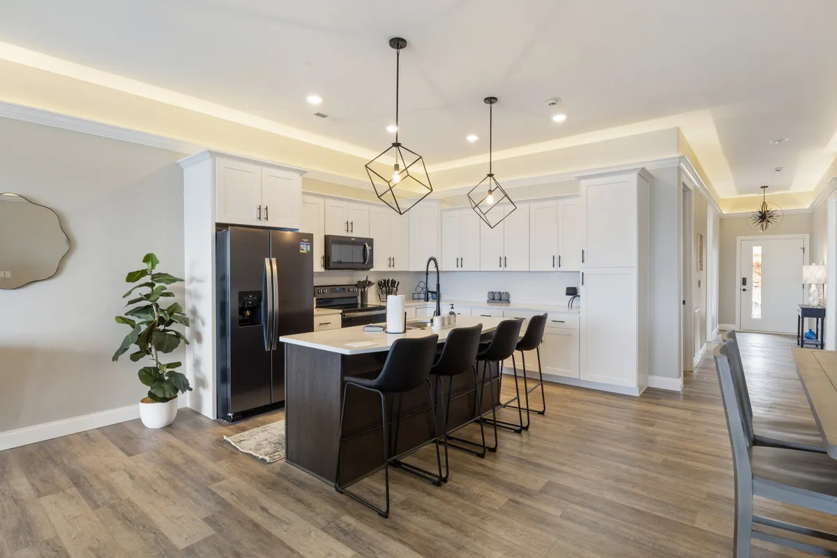 A stylish kitchen with a prominent island, bar stools, pendant lights overhead, and appliances in the background. Greenery and decor elements add a homelike feel, while the space appears organized and ready for both cooking and entertaining.