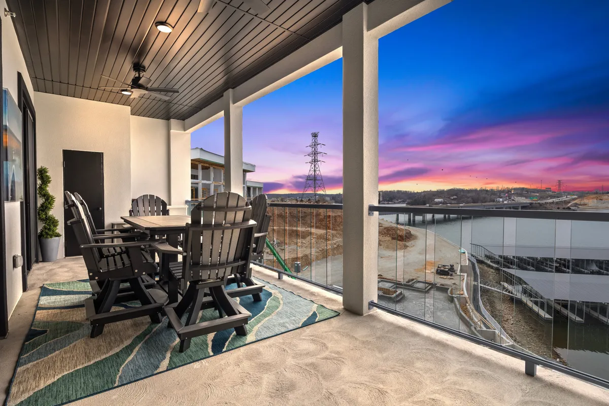A balcony scene showcasing a dining table with chairs, a decorative rug, and a sweeping, scenic view of the water at sunset. The space is designed for socializing and unwinding, capturing the magic of lakeside evenings and the feeling of a private retreat.