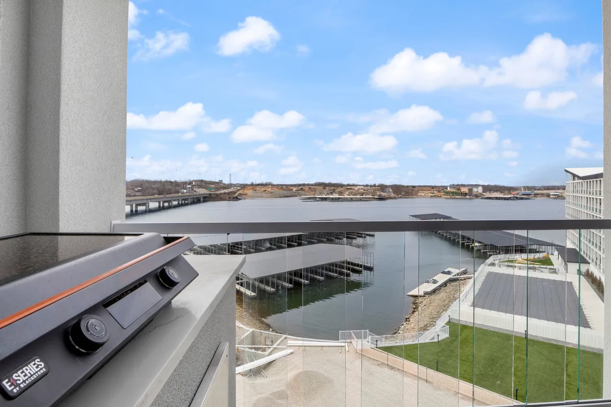 A view from a private balcony featuring a sturdy railing, an electric grill set up for barbecuing, comfortable outdoor seating, and a panoramic view over the water and dock area. The scene suggests relaxation and social gatherings, with boats visible in the distance and the sky hinting at a serene time of day.