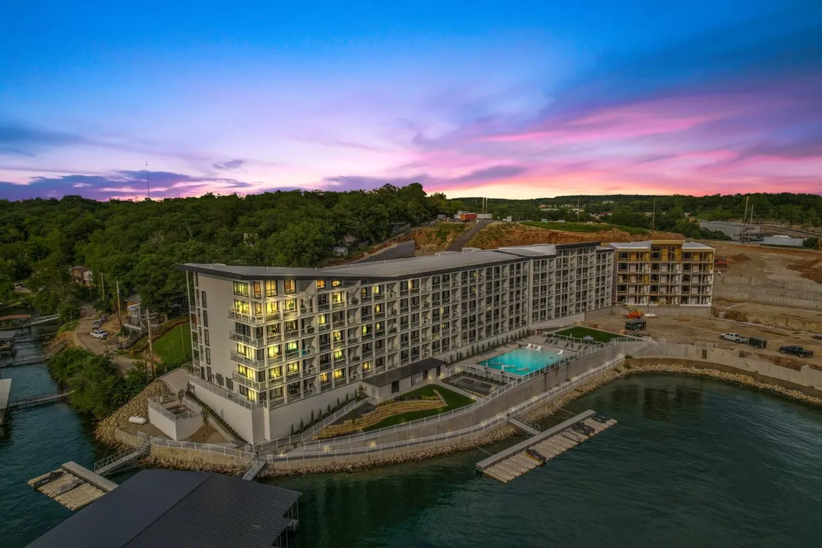 This exterior image features a modern hotel building set against a vibrant sunset sky, a swimming pool, a waterfront dock, and lush greenery. Guests can be seen enjoying serene moments by the water, as well as socializing by the pool, reflecting the lively yet relaxing spirit of Osage Beach.