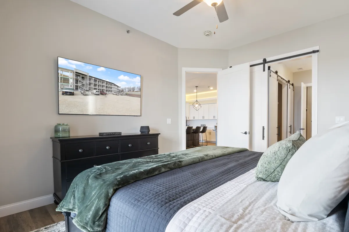 A stylish bedroom with a bed featuring a green throw blanket, a black dresser, and a television displaying an exterior view. The decor is modern and comfortable, with a focus on relaxation and entertainment.