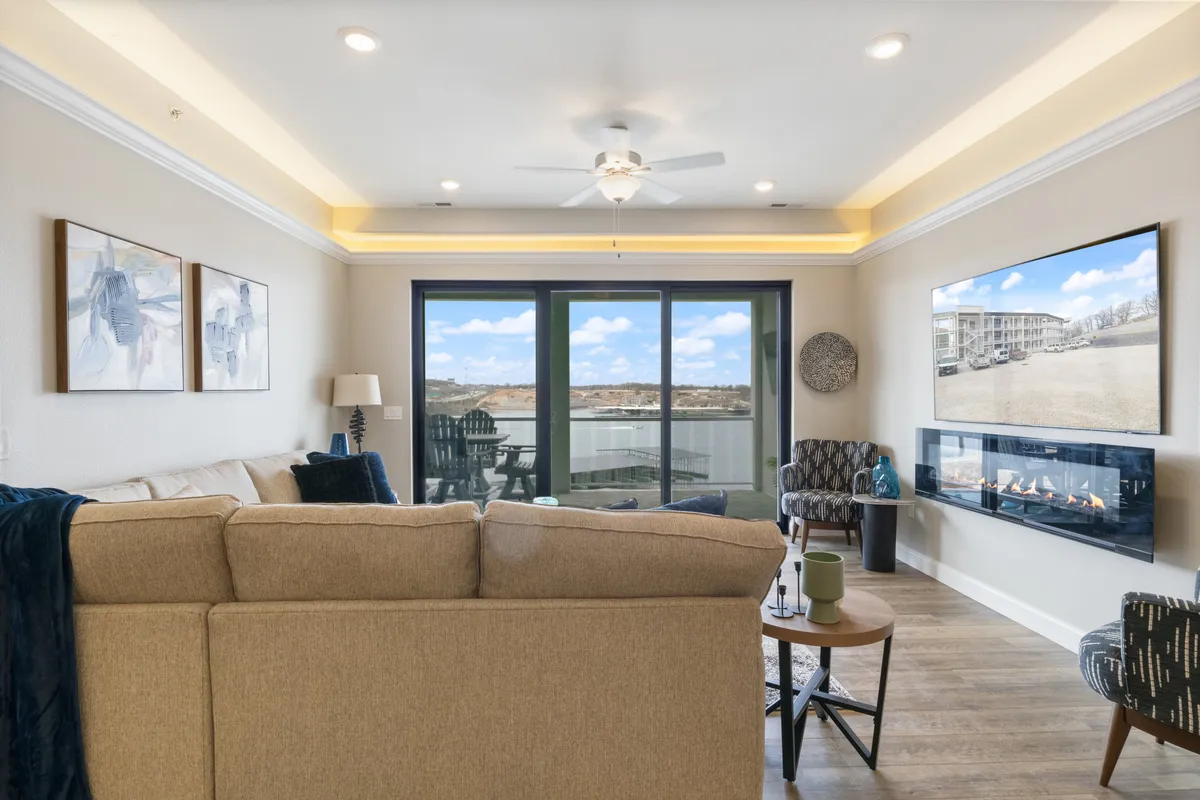 This image showcases a cozy living area with a plush sofa, an electric fireplace, a smart TV, and large windows that offer expansive views of the lake. The space is bathed in natural light, and tasteful artwork further enhances the inviting atmosphere.