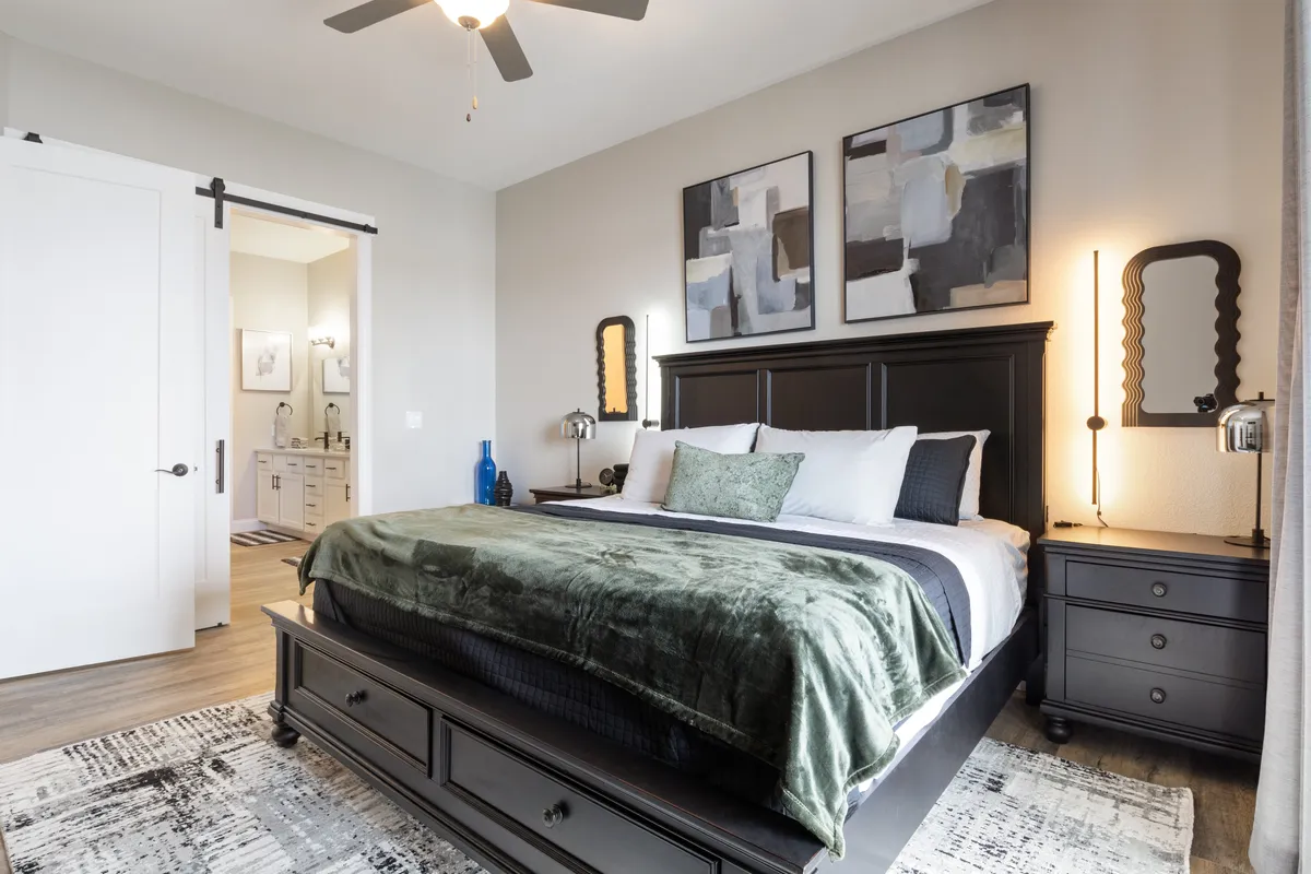 A calm and sophisticated bedroom featuring a king-sized bed with a dark wooden frame, accented by a green throw blanket and abstract artworks above the headboard. Mirrored wall sconces add ambient lighting, and large windows allow natural light to filter in, creating an inviting retreat.