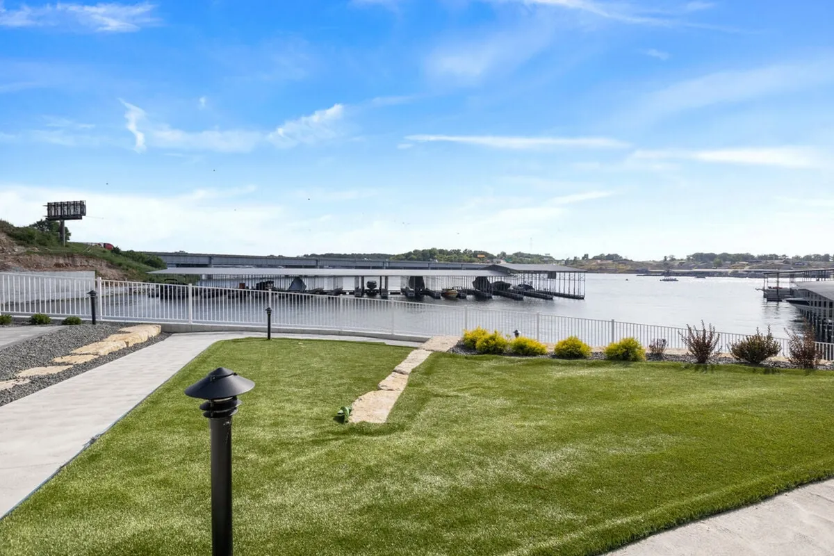 A tranquil waterfront scene features a green lawn leading to the edge of Lake of the Ozarks. Several boats are docked at a nearby pier, and the area is bordered by a modest fence. The calm water and natural setting evoke an inviting sense of relaxation and outdoor recreation.