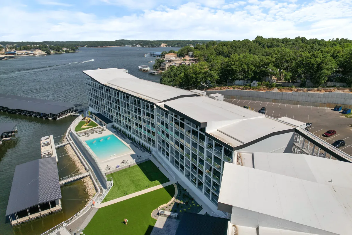 This exterior image captures the modern building structure with a large swimming pool in the foreground, a green lawn area, and the waterfront just beyond. The pool deck looks inviting for summer activities, while the expansive view of the water hints at boating and lake adventures.